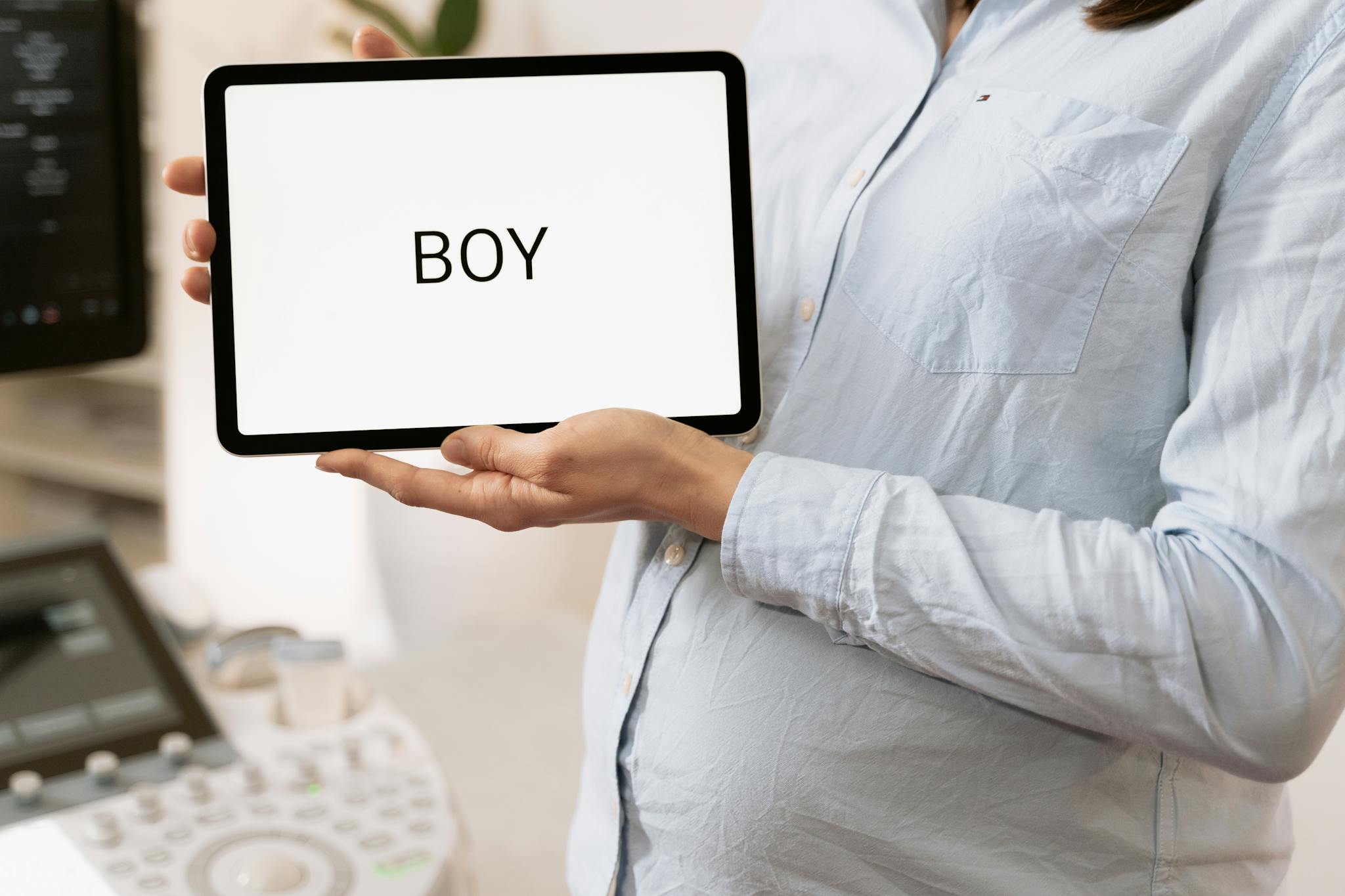 Pregnant woman holding a gender reveal tablet display in a medical setting.